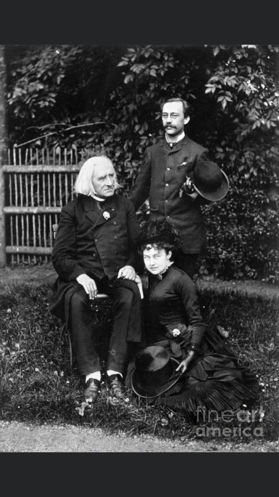 Picture in black and white taken in 1884, showing Liszt sitting in his garden, wearing a black coat. His pupil Carl Lachmund is standing behind him, and Caroline Lachmund, Carl's wife, is sitting on the grass on Liszt's right. Liszt is holding Caroline's hand. Carl and Caroline wear black jackets and hold their hats.