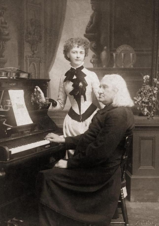 Picture in black and white taken in 1885. It represents Liszt and his student Arma Senkrah. Liszt is sitting at an upright piano. He wears a cassock. One of his hands is on the keyboard. A score of Brahms is on the music stand. Arma stands behind him. She wears a white dress with a black ribbon. Her violin is on the upper part of the piano.