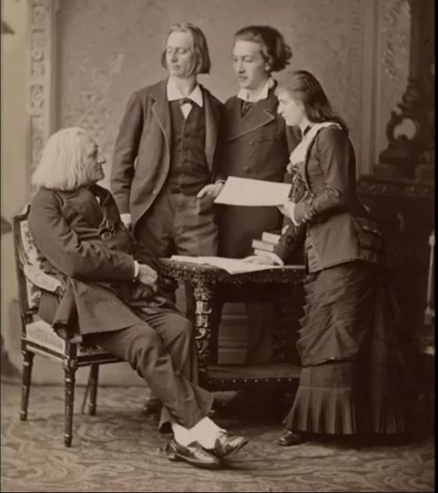 Picture in black and white taken in 1881. It represents Liszt surrounded by his students Juliusz Zarebski, Franz Servais and Johanna Wenzel. Liszt is sitting in an armchair and looks at his students who are standing behind him, looking at Liszt. Johanna has a score in her hand.