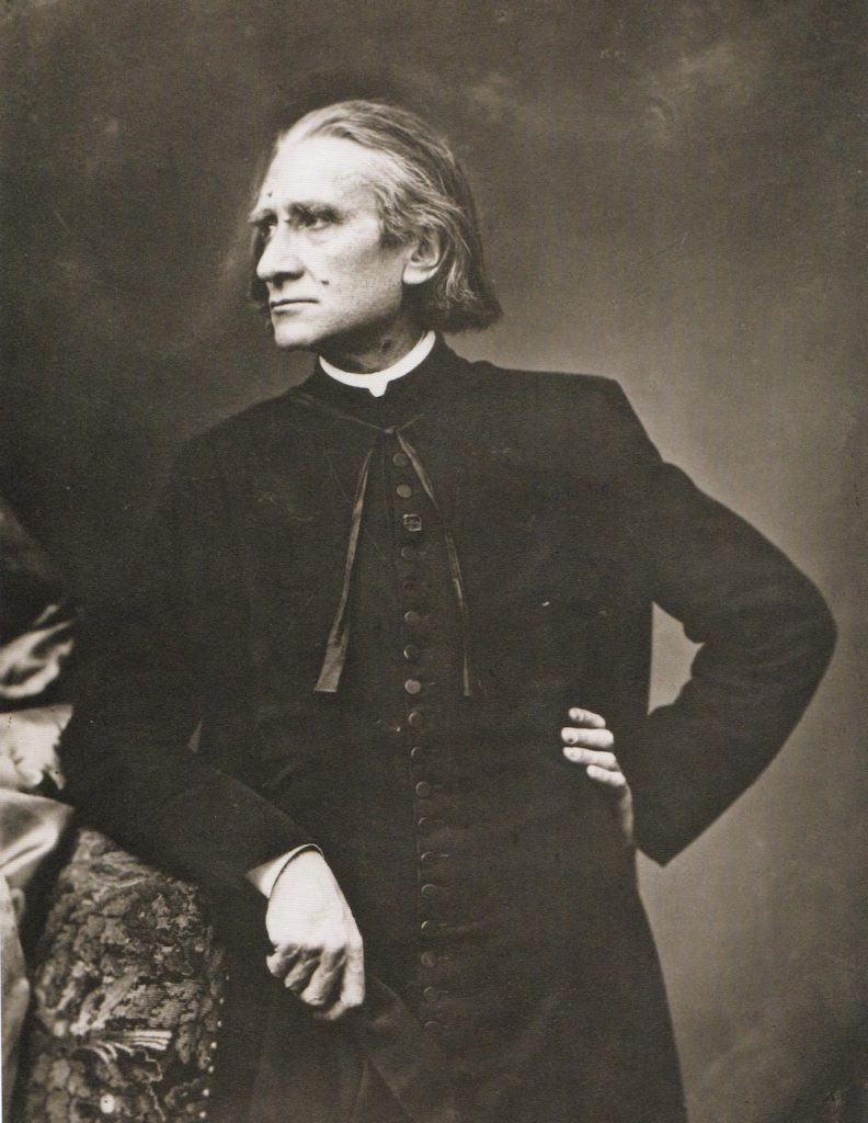 Picture in black and white taken in 1866. Liszt wears a cassock. His head is turned to the right. His left hand is on his hip and his right arm leans on a table.