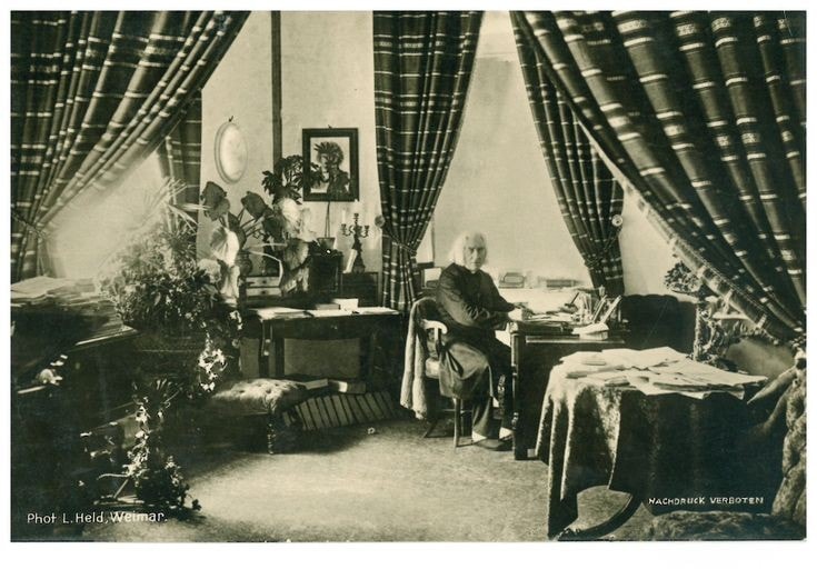 Picture in black and white taken in 1884. It represents Liszt's living room in Weimar. A part of his grand piano can be seen on the left. There are large curtains at the windows, a portrait of Beethoven on a wall on the left, two desks, plants, many books and scores on a table and on the piano. Liszt is sitting at a desk on the right and looks at the camera. He wears a black cassock. He has a pen in his right hand and seems to write something on a paper.