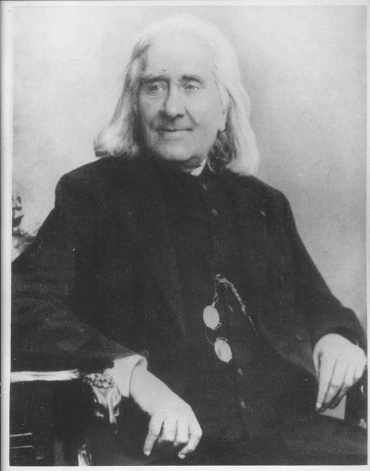 Picture in black and white taken in 1886. It represents Liszt sitting on an armchair. He wears a cassock. His glasses are attached to one of the buttons. He is facing the camera and smiling.