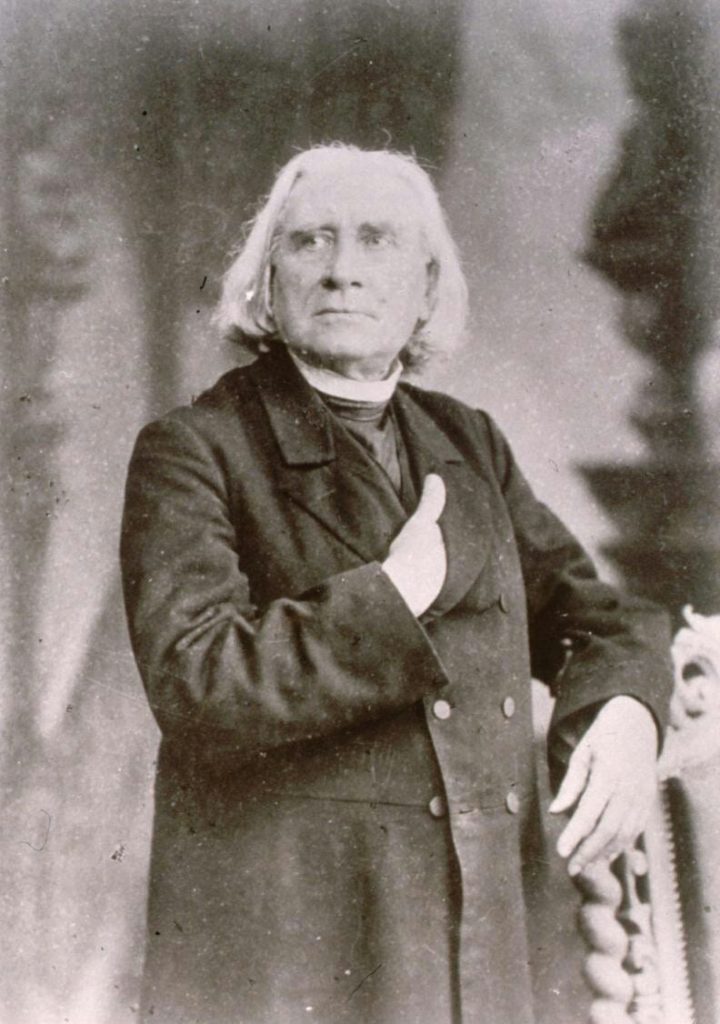 Picture in black and white taken in 1883. It represents Liszt standing, wearing a black cassock and a black coat. His right hand is inside his coat. His left arm rests on the back of a chair. He looks toward his right.