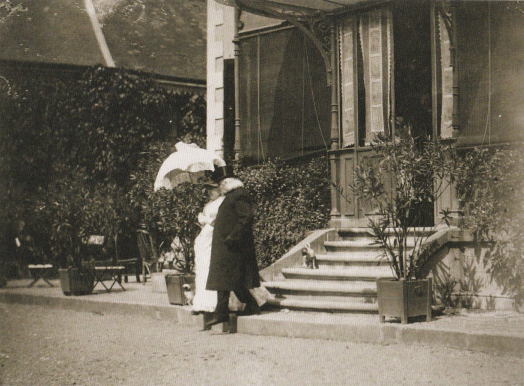 Picture in black and white taken in 1886. It represents Liszt and  Cécile Munkácsy walking down the stairs of a house, toward a garden. Cécile is wearing a white coat and an umbrella. Liszt is wearing a black coat and a hat.