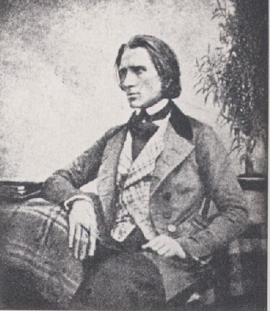 Picture in black and white taken in 1846. It represents Liszt sitting on a chair in front of a table. He looks toward his right. His right arm leans on the table. His left hand is on his leg. He wars a grey coat over a checked jacket. The tablecloth is also checked.