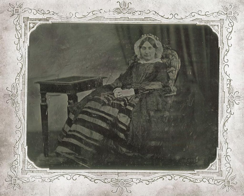 Picture in black and white taken in 1847. It represents Liszt's companion, Princess Carolyne zu Sayn-Wittgenstein, sitting in an armchair in front of a table. She wears a stripped grey and black dress and has a white bonnet on her head. She faces the camera and smiles.