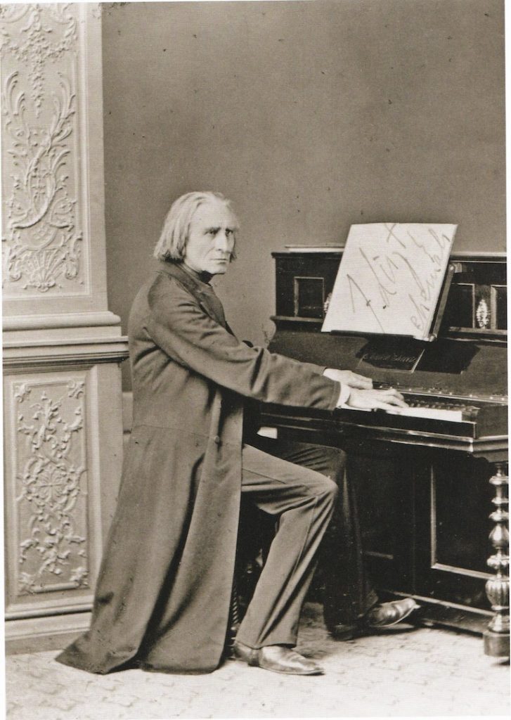 Picture in black and white taken in 1869. It represents Liszt in front of an upright piano. He wears a black long coat and black pants. His hands are on the keyboard. He looks toward the camera.