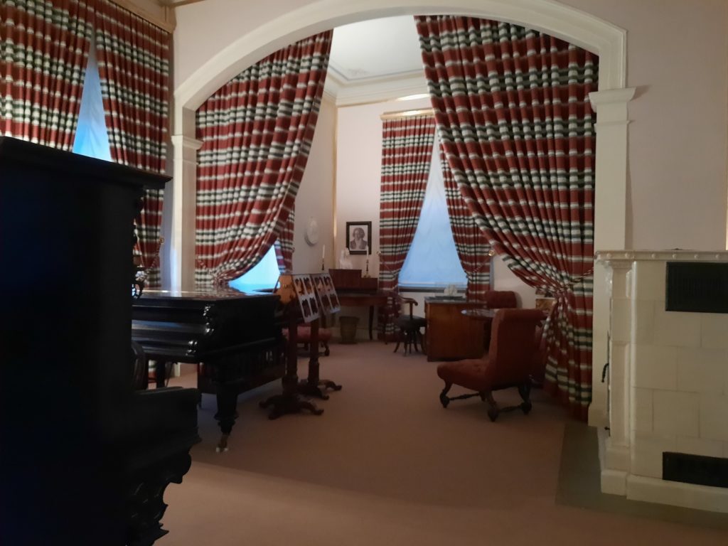 Picture in color taken in 2021. It represents the interior of Liszt's living room in Weimar. There are large red curtains on the windows. On the left we can see part of an upright piano, and a grand piano. There are two old music stands close to the grand piano. On the right, a small table with two brown chairs. In the back, a portrait of Beethoven on the left wall, and a desk on the right.