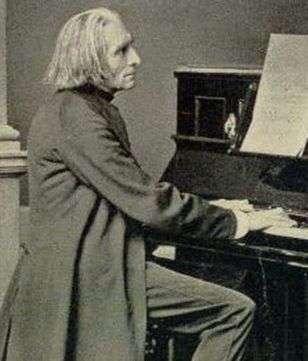 Picture in black and white taken in 1869. It represents Liszt in front of an upright piano. He wears a black long coat and black pants. His hands are on the keyboard. He looks at a score.