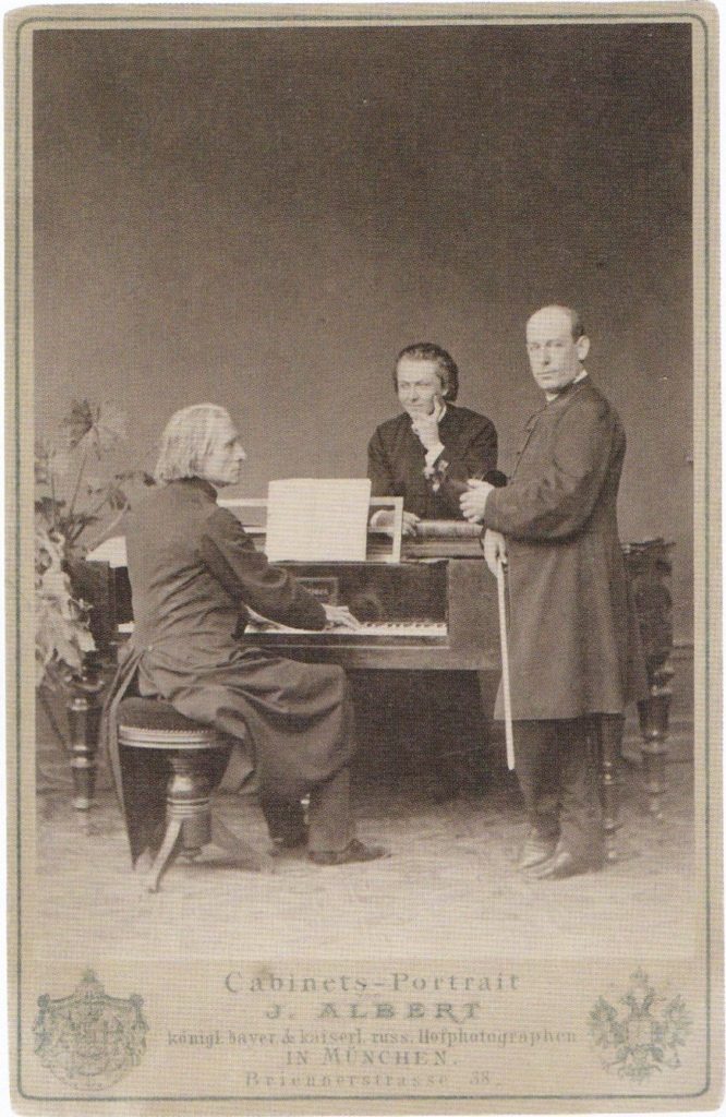 Picture in black and white taken in 1867. It represents Liszt sitting in front of a piano, wearing a black coat. His students Ede Reményi and Nándor Plotényi stand behind the piano. At the bottom of the picture, the name of the photographer and his address.