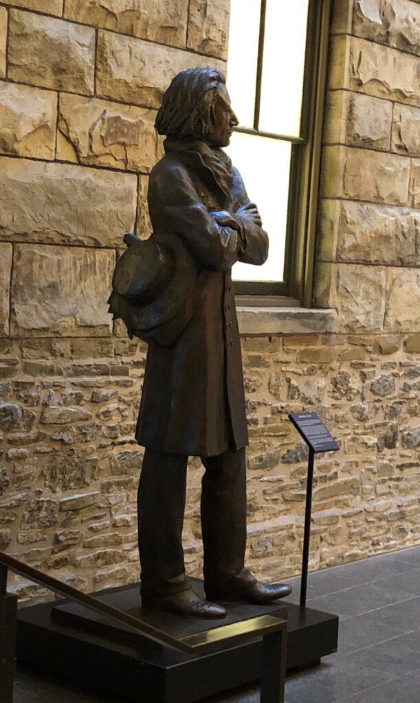 Liszt's bronze statue seen from the back. We can see him standing, arms crossed, and holding a hat with his right hand.