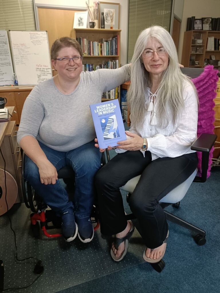 Diane and Evelyn, two white women, smile at the camera. Diane sits on a wheelchair, she has short brown hair, she wears glasses, a grey top and a jeans. Evelyn has long white hair, wears glasses, a white top and a black pant. Evelyn holds the book entitles "Women's Leadership in Music." 