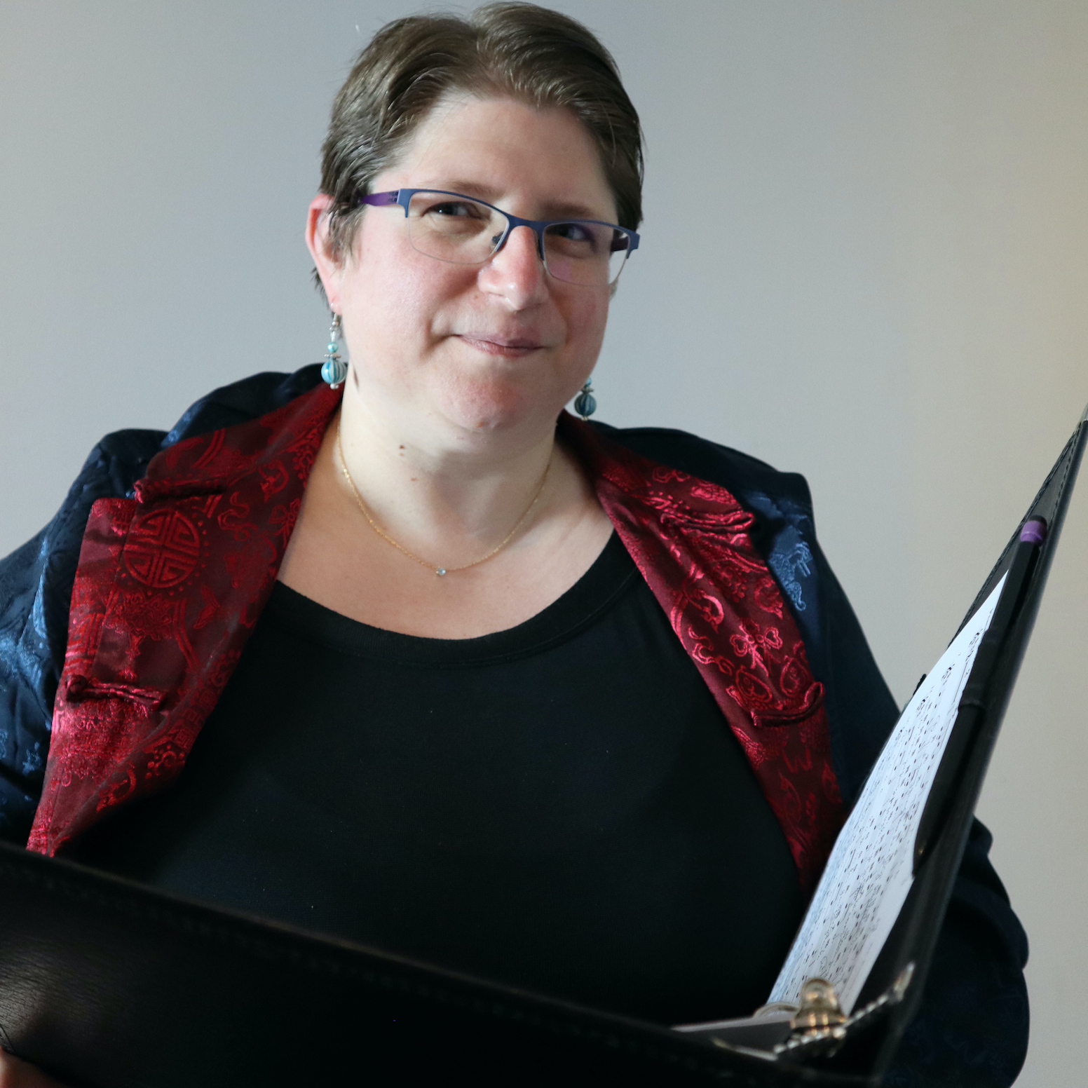 A white woman with short brown hair wearing glasses, earrings, a blue and red vest, and holding a folio.