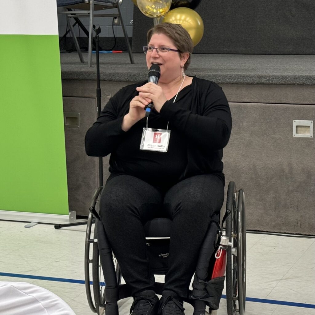 Diane in her wheelchair is dressed with black suit a black pants. She talks to an audience using a microphone.