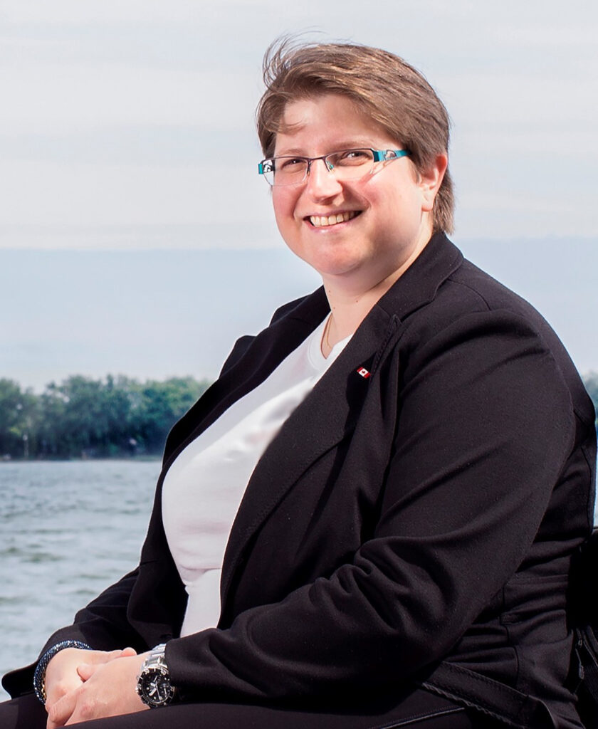 Diane, a woman with short brown hair and blue framed glasses, wearing a white t-short and a black jacket with a pin representing a Canadian flag. She smiles at the camera.