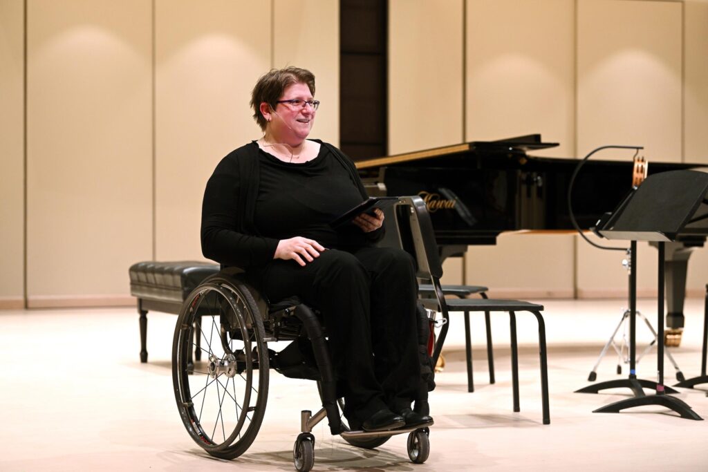 Diane in her wheelchair dressed in her black presentation suit, she has a micro-headset and smiles at the audience while giving a keynote.