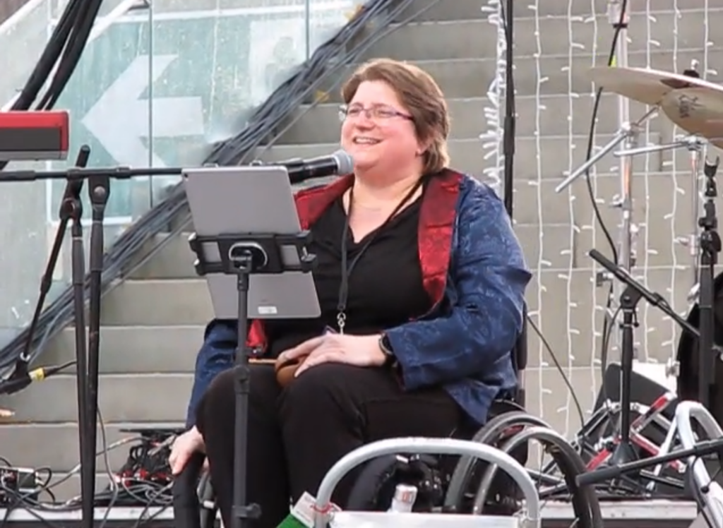 Diane in her wheelchair singing in a microphone on a stage surrounded by instruments, she smiles while singing.