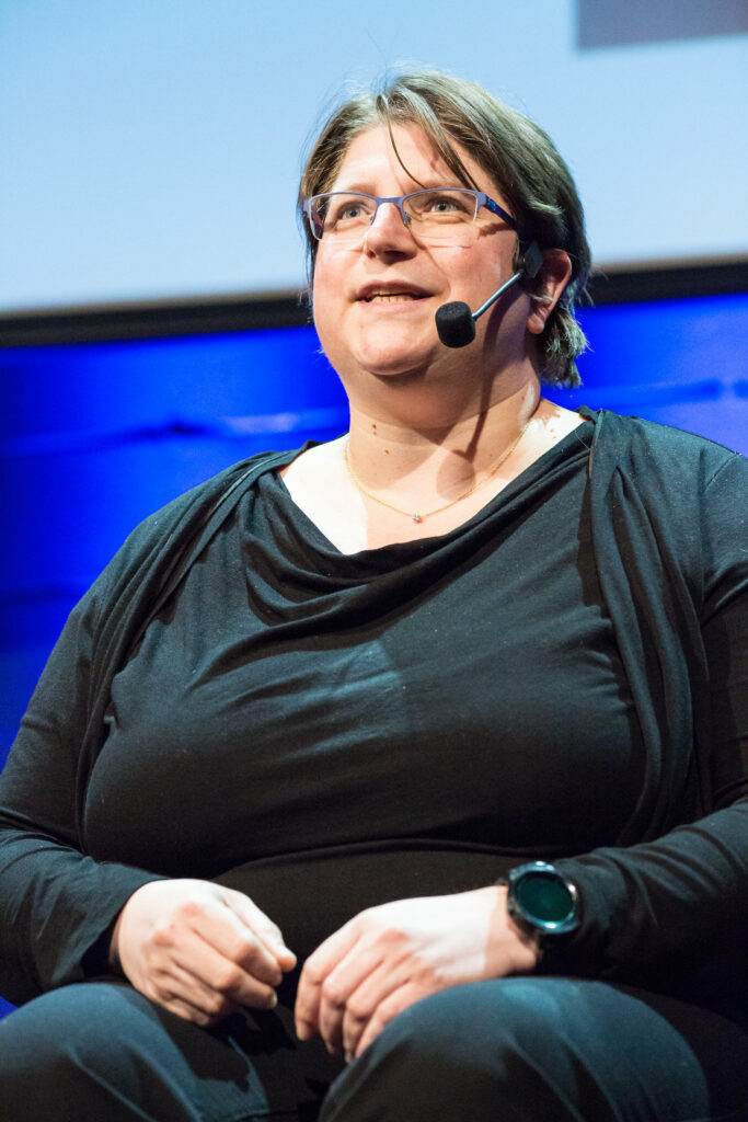 Diane, a white woman with short brown hair, blue eyes, and glasses, wears a black outfit. She speaks to an audience through a micro-headset.