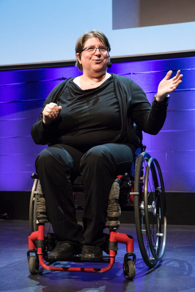 Diane sits in her wheelchair, she wears a black costume. She has short brown hair, glasses, and a micro-headset. She speaks to the audience, moving her arms, and smiling.