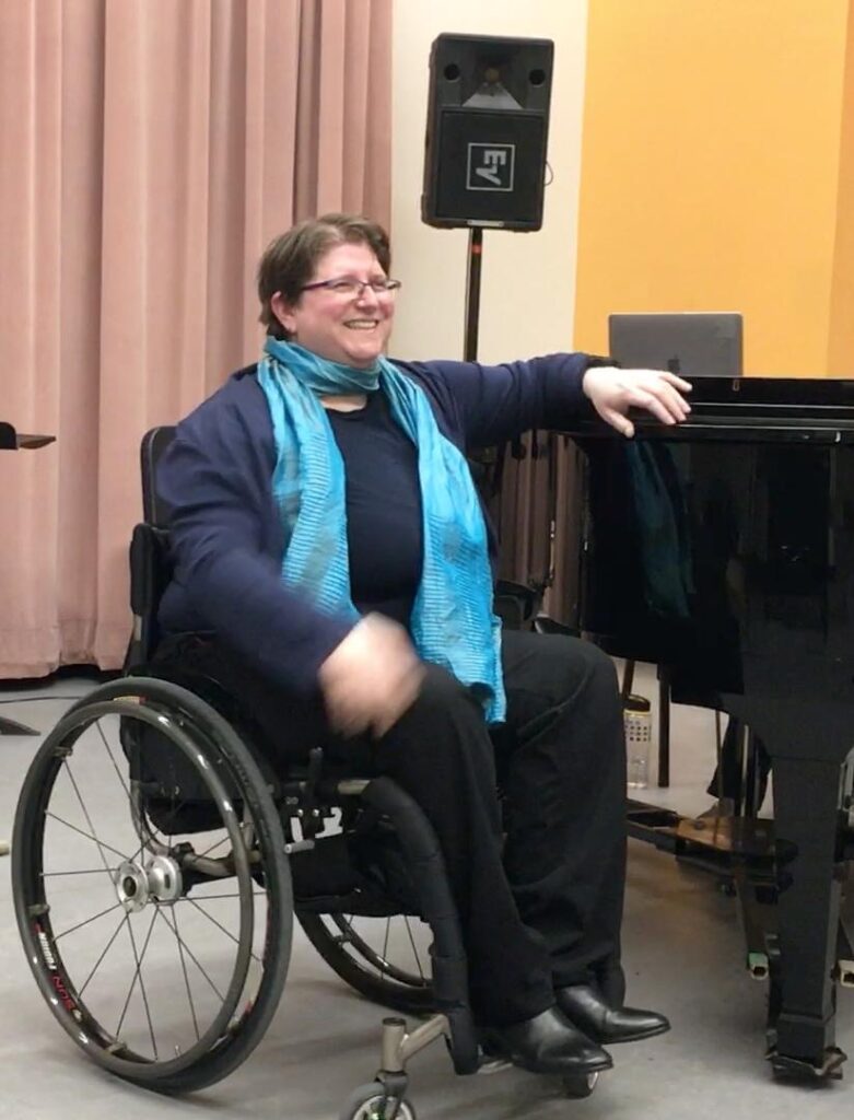 Diane in her wheelchair wearing a blue ensemble and a green scarf, she smiles at the audience while singing, her left arm is on a black grand piano.
